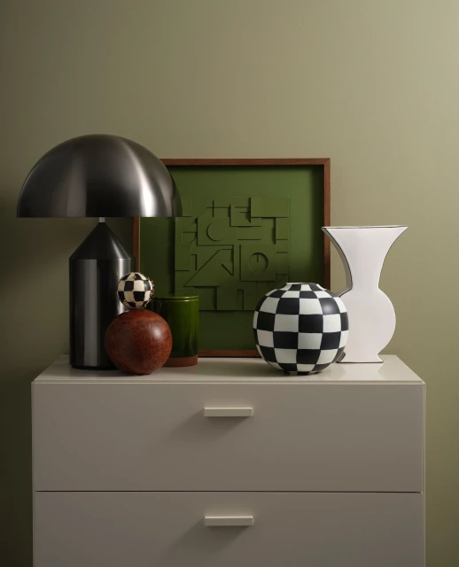 Still life composition featuring a lamp, vases, and framed art on a white dresser against an olive green wall in an indoor setting.