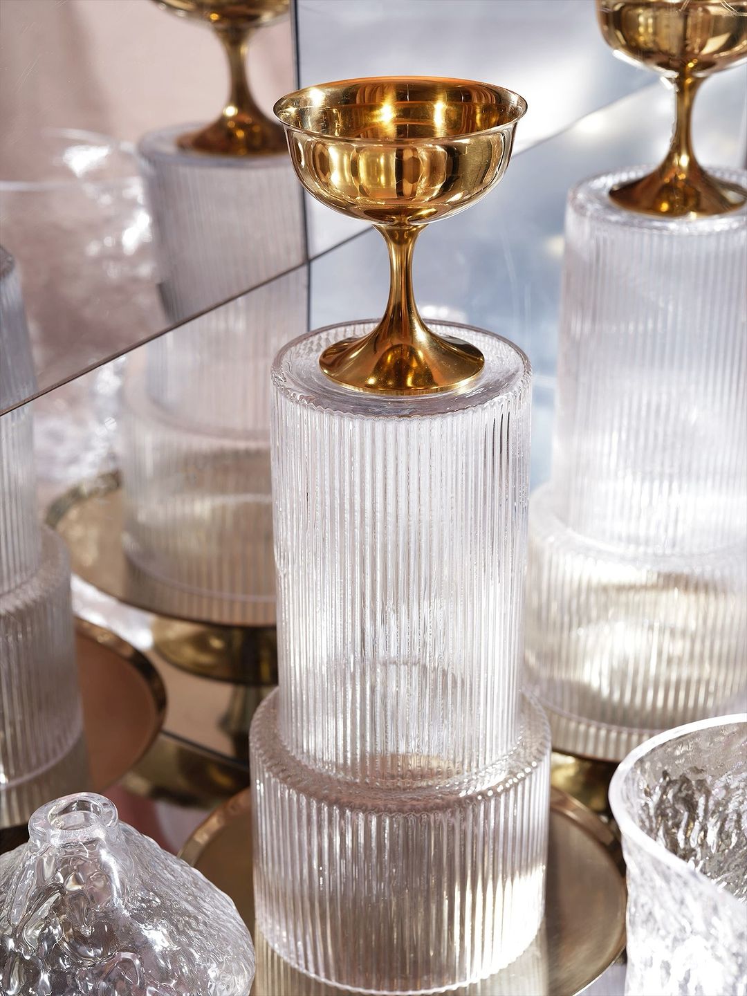 Golden coupe glass stands atop clear ridged pedestals reflecting in a mirror. Shiny textures, studio setting, neutral color palette.
