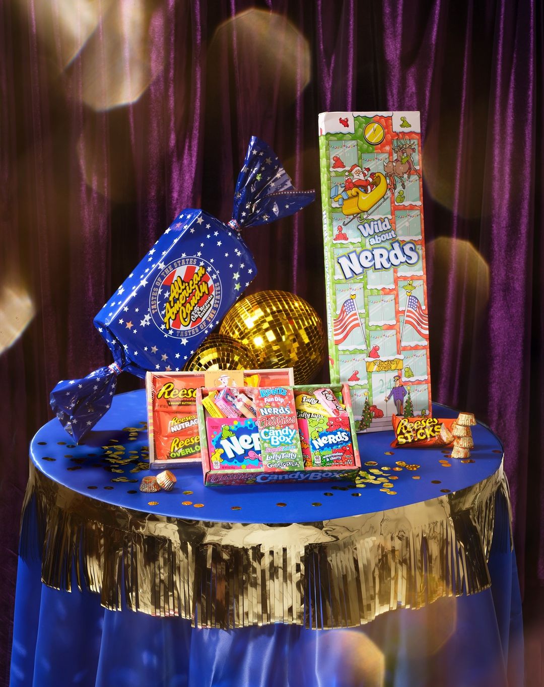 A collection of colorful candy boxes, a disco ball, and confetti are arranged on a blue tablecloth with gold fringe.