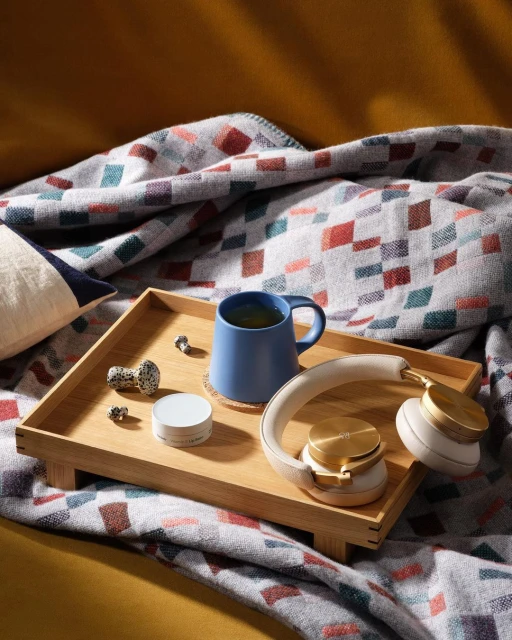 Still life shot of a blue ceramic mug with tea, a pair of wireless headphones, and small decorative objects on a wooden tray atop a blanket.