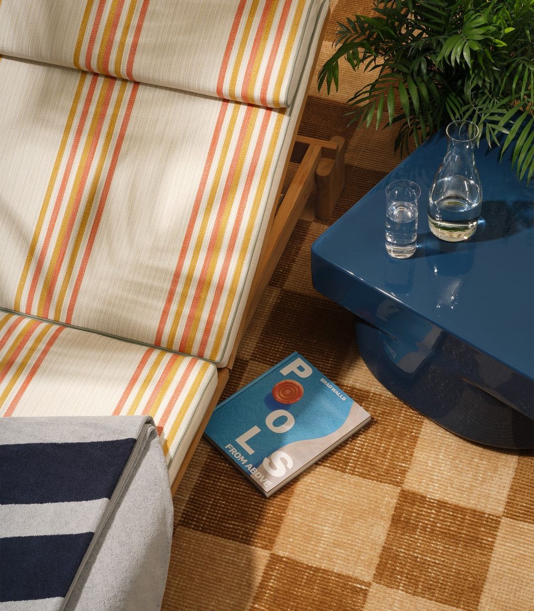 Overhead shot of striped lounge chair, turquoise table with glass of water and carafe, a book titled 'Pools From Above', and potted plants.