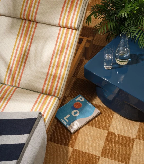 Overhead shot of striped lounge chair, turquoise table with glass of water and carafe, a book titled 'Pools From Above', and potted plants.