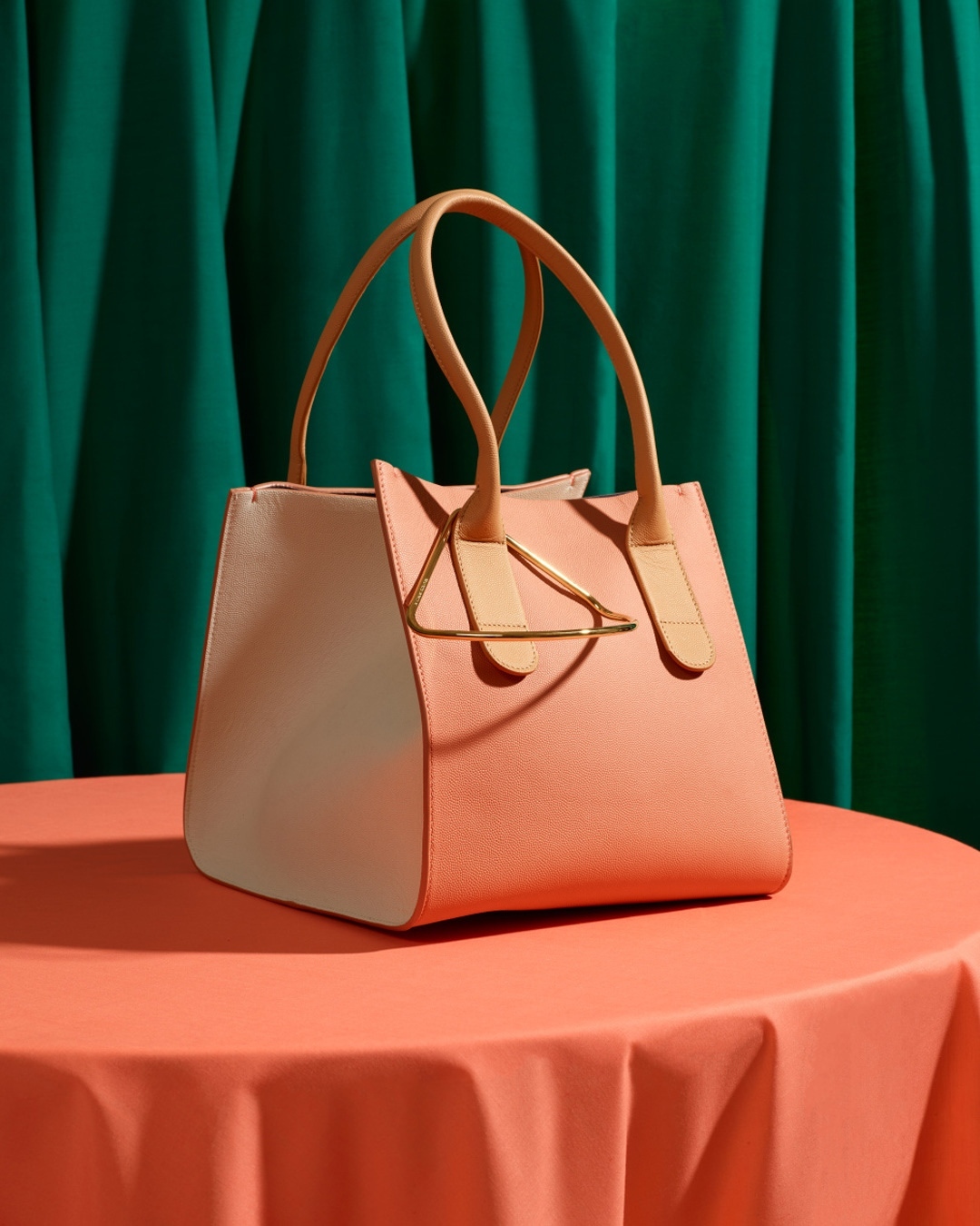Elegant pink and white leather handbag with beige handles and a golden triangular accent piece on a peach-colored draped table.