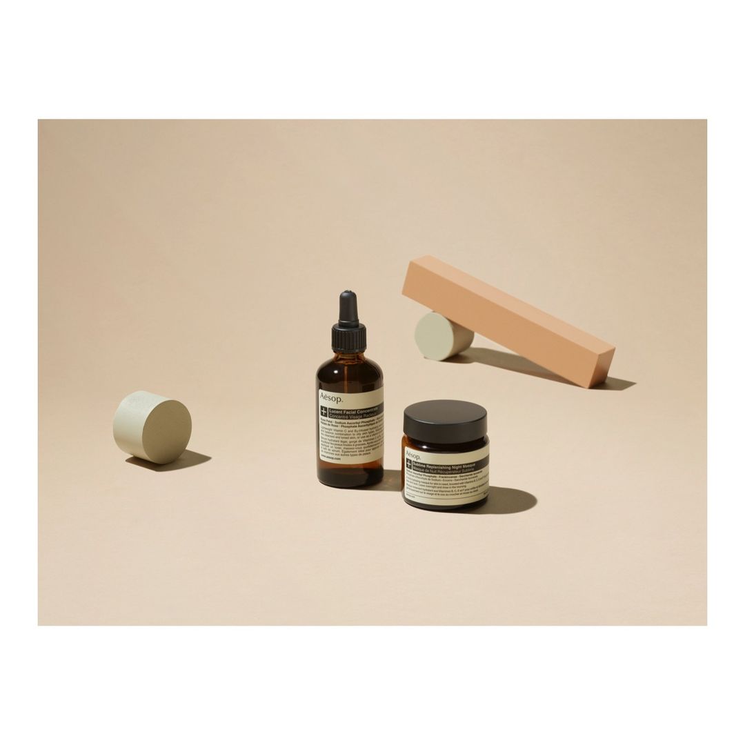 Two amber glass bottles of Aesop skincare products are featured alongside beige geometric shapes against a light beige background.