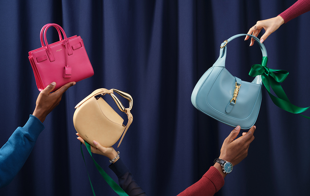 Three luxury purses, including Saint Laurent and Chloé, are displayed by two people against a dark blue studio backdrop.