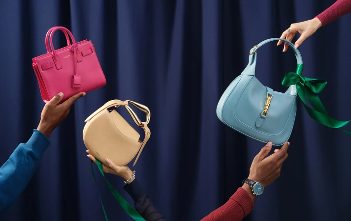 Three luxury purses, including Saint Laurent and Chloé, are displayed by two people against a dark blue studio backdrop.