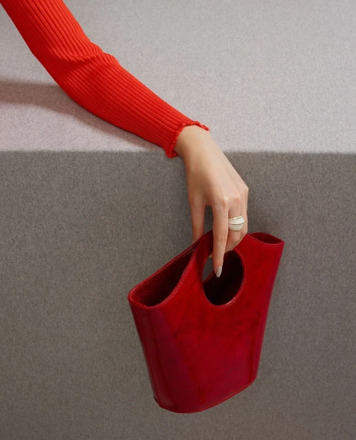 Close-up shot of a woman's hand holding a glossy red handbag against a gray textured background, focusing on fashion accessory.