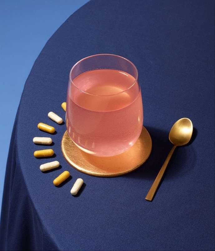 Close-up of pink drink in clear glass on gold coaster surrounded by supplement pills and a gold spoon on a blue tablecloth.