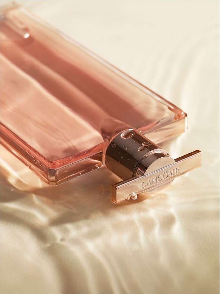 Close-up studio shot of a Lancôme perfume bottle with a rose-gold cap, sitting on a shimmering surface with light reflecting like water.