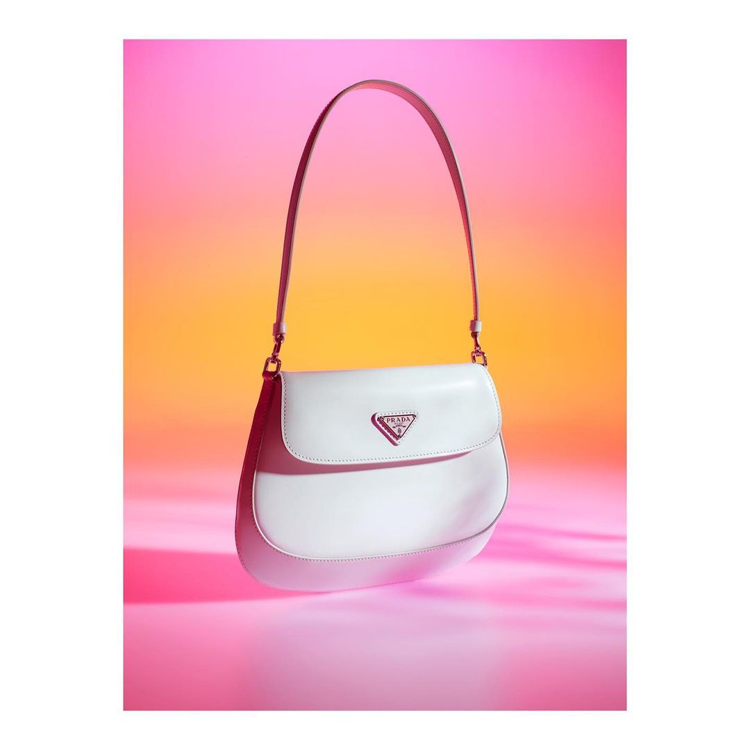 White Prada handbag with pink logo against orange and pink gradient background. Fashion accessory for luxury brand marketing and retail.