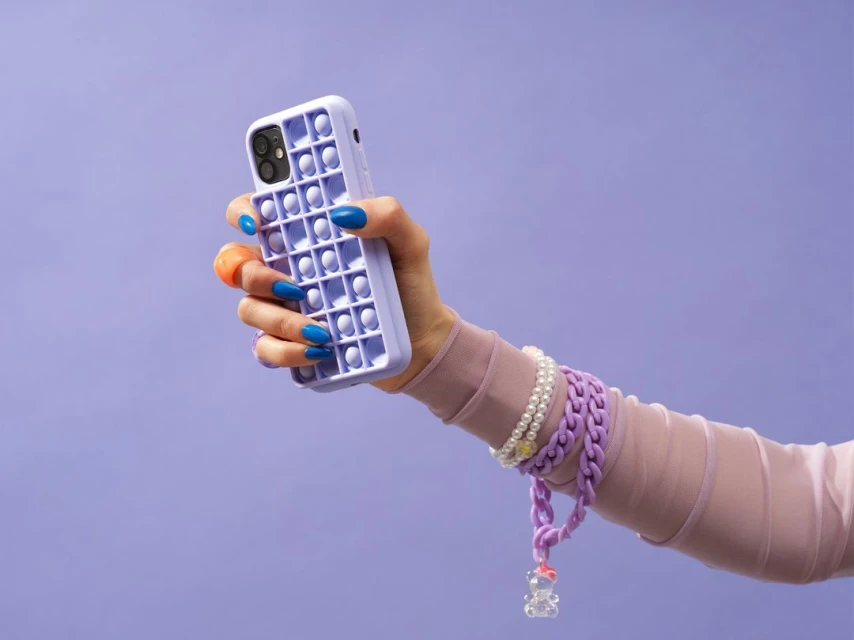 Hand holds a lavender phone case with a pop-it design, accessorized with bracelets and set against a light purple backdrop.