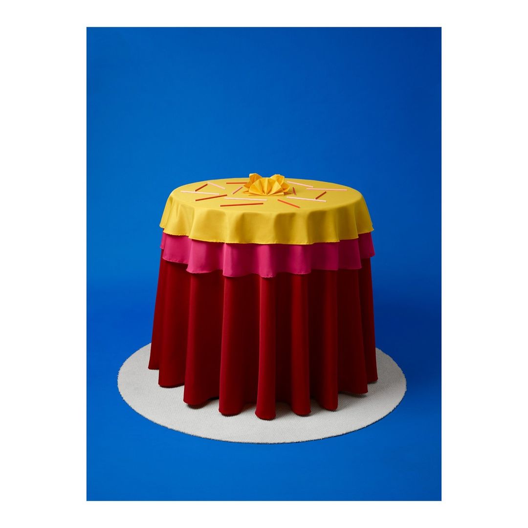 Three-tiered round table covered in yellow, pink, and red fabric with yellow flower accent on a white platform against a blue backdrop.