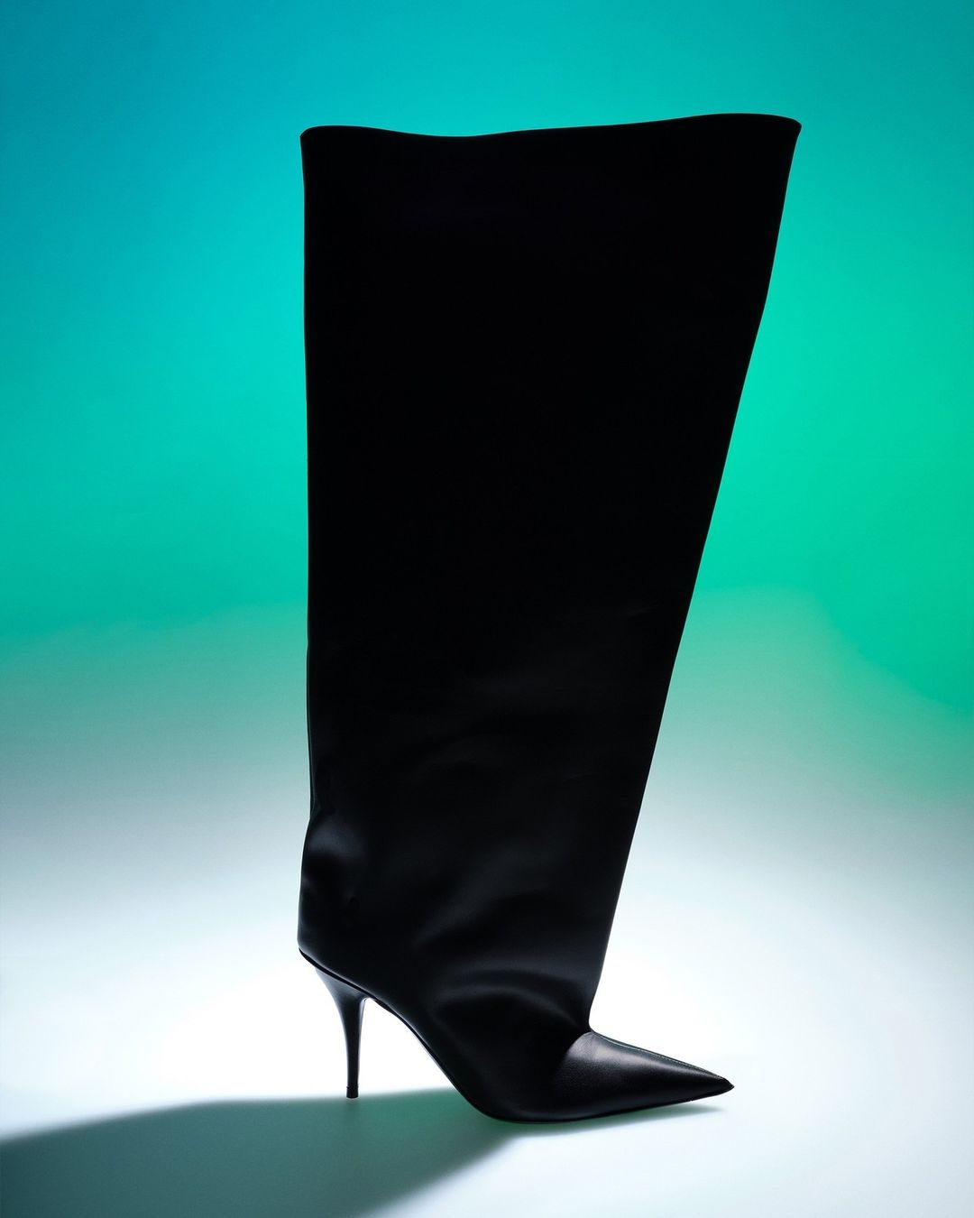 Black leather over-the-knee stiletto heel boot with a pointed toe on a teal and blue gradient background in a studio setting.