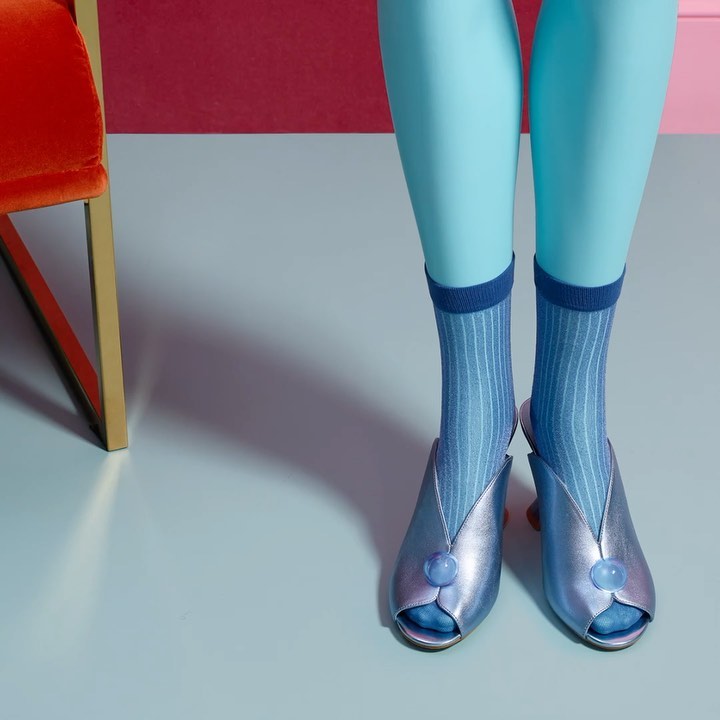 Turquoise legs wearing blue striped socks and light purple metallic high heels are positioned on a gradient studio backdrop.