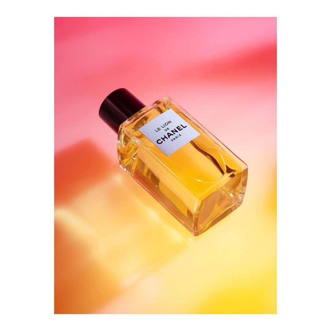 Close-up of a rectangular clear glass Chanel perfume bottle filled with golden liquid, against a pink and yellow gradient background.