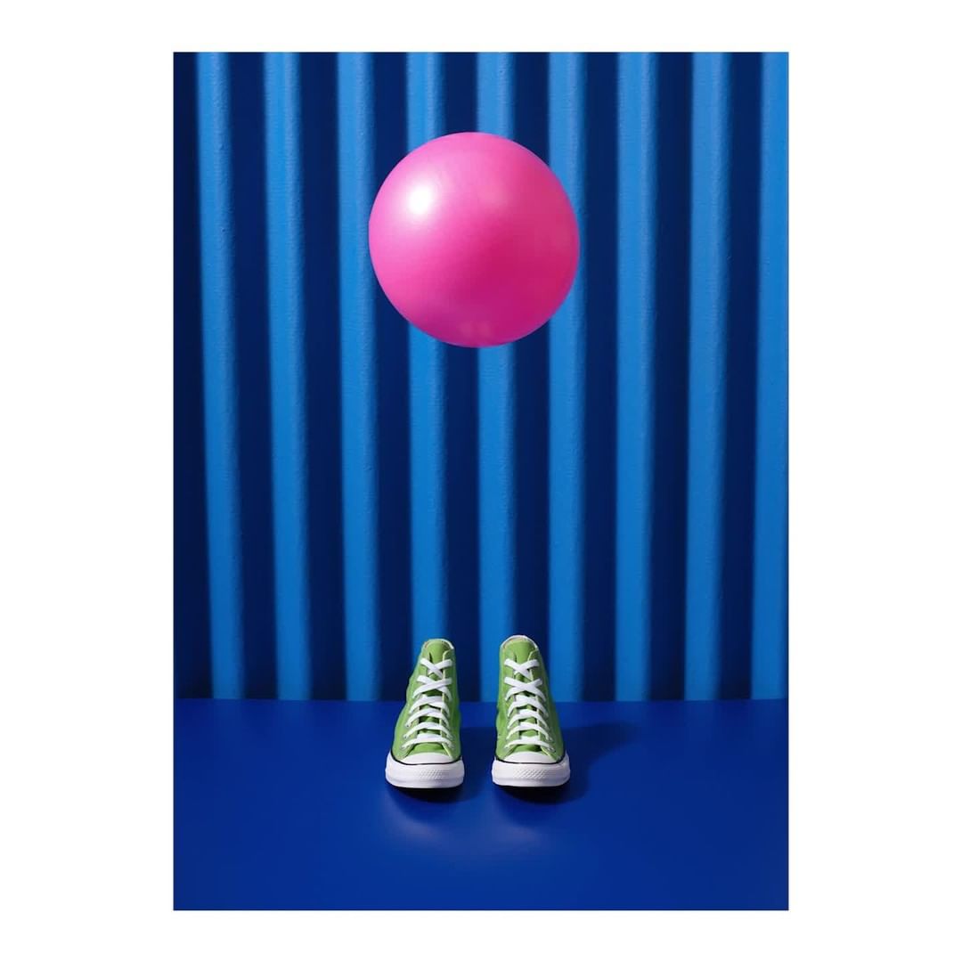 Studio shot featuring green high-top sneakers on a blue surface, contrasted by a pink ball against a blue striped background.