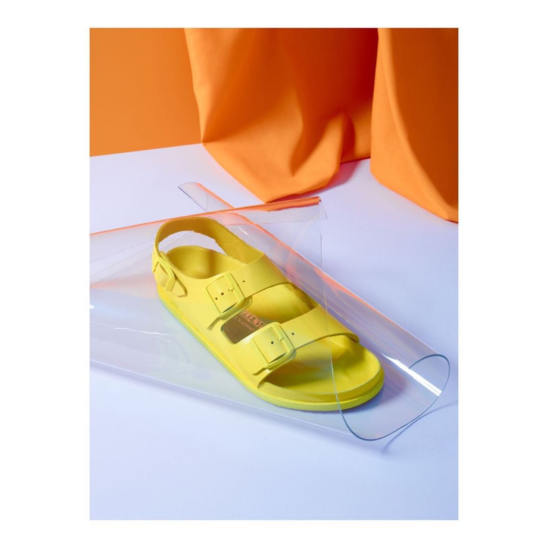 Yellow Birkenstock sandal on a clear surface with orange background. The photo features the footwear on a transparent sheet.