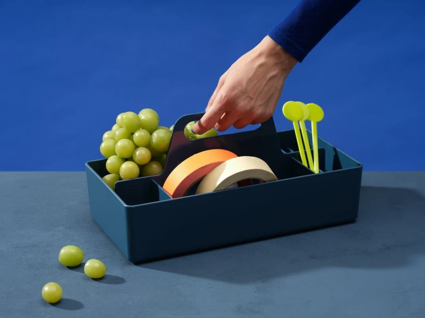 Still life of grapes, tape rolls, and yellow sticks in a blue carrier, held by a person's hand, set against a blue background.