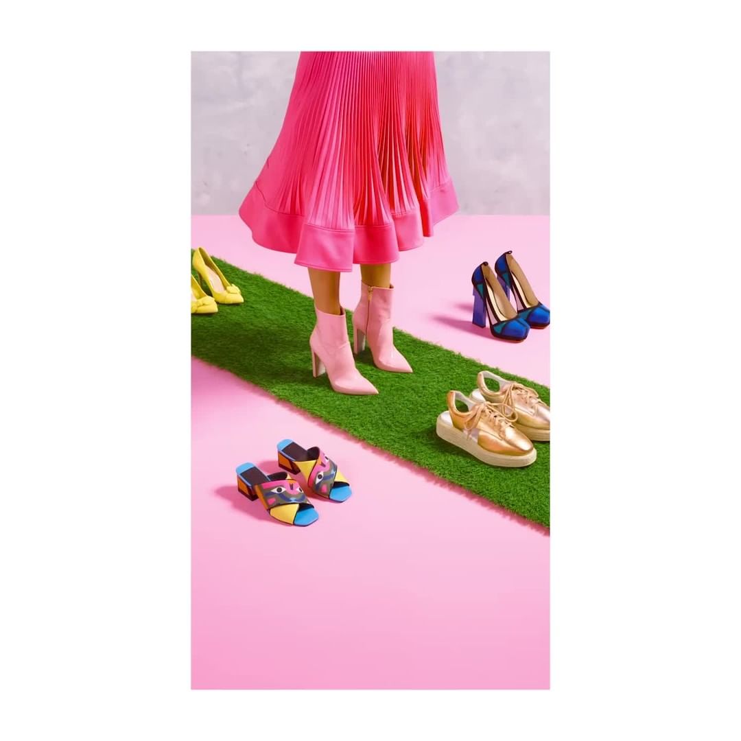 Bright, colourful image of a person wearing a pink pleated skirt and pink boots standing on a strip of artificial grass surrounded by shoes.
