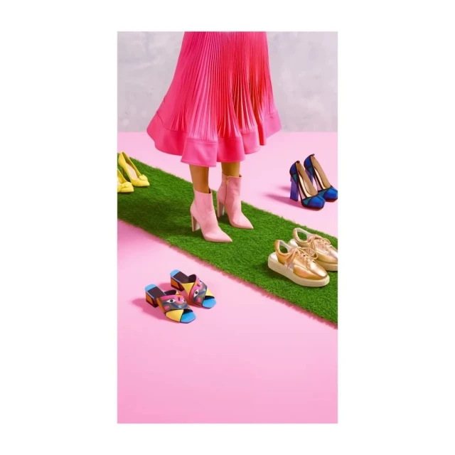 Bright, colourful image of a person wearing a pink pleated skirt and pink boots standing on a strip of artificial grass surrounded by shoes.