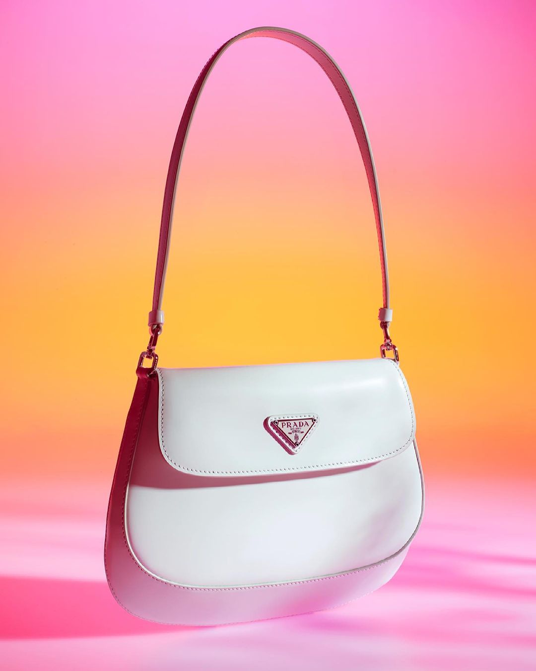White Prada handbag with a curved shape and handle, displayed against a vibrant gradient background of pink and orange hues.