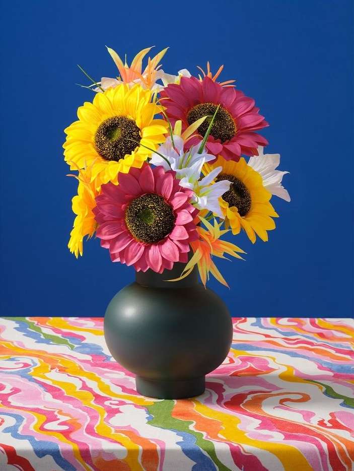 Floral arrangement with yellow and pink sunflowers and white lilies in a dark green vase on a patterned tablecloth, against a blue background.