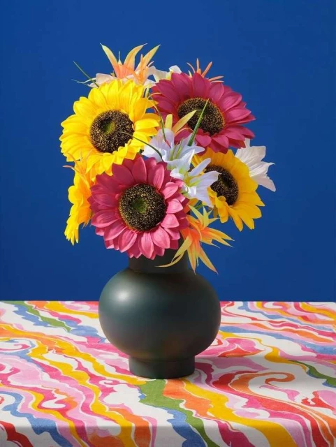 Floral arrangement with yellow and pink sunflowers and white lilies in a dark green vase on a patterned tablecloth, against a blue background.
