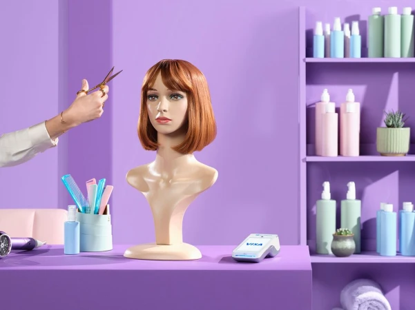 Hair salon scene with stylist's hand holding scissors near wig head, combs, product bottles, and POS system on violet surface.