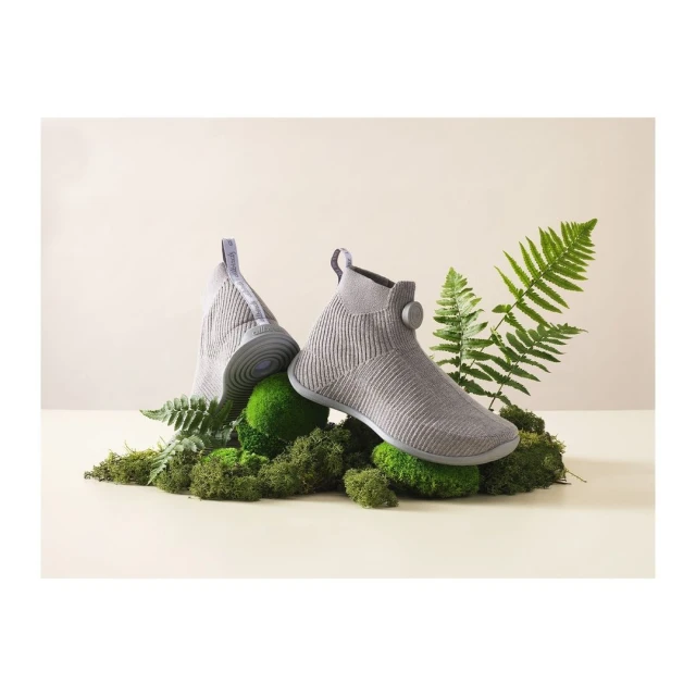 Two gray knitted shoes on moss and ferns against a cream background. The shoes are elevated on a surface. Studio lighting.