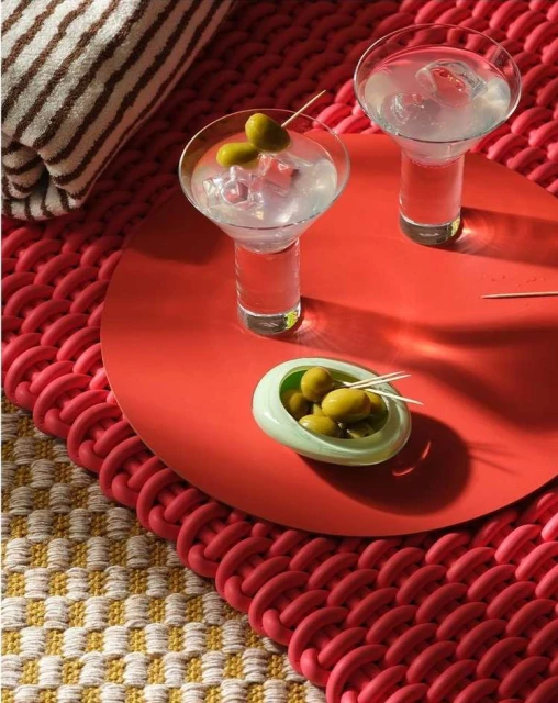 Two martini glasses with green olives and ice sit on a red tray with a striped towel and woven mat in the background.