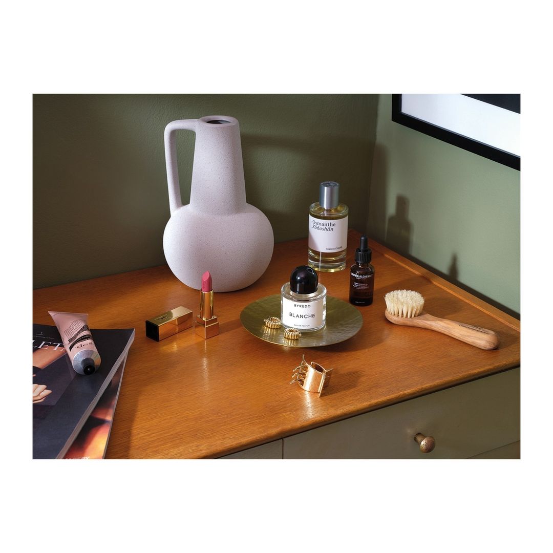 Cosmetic arrangement on a wooden dresser features perfume bottles, lipstick, lotion, jewelry, a hairbrush, and a fashion magazine.