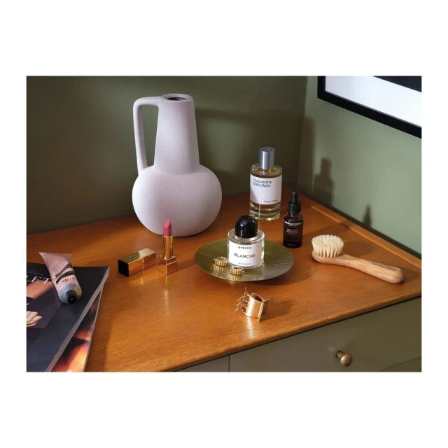 Cosmetic arrangement on a wooden dresser features perfume bottles, lipstick, lotion, jewelry, a hairbrush, and a fashion magazine.