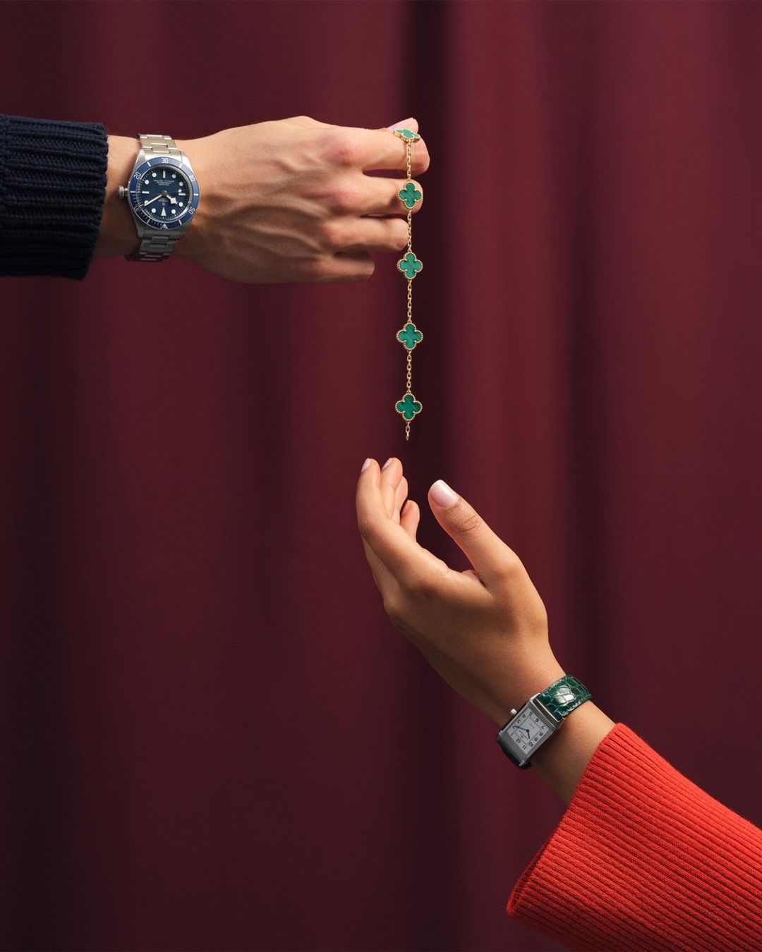 Close-up of two hands, one holding a gold necklace with green clover-shaped stones, against a maroon background, jewelry and watches visible.