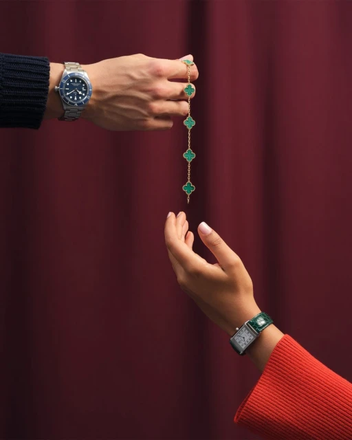 Close-up of two hands, one holding a gold necklace with green clover-shaped stones, against a maroon background, jewelry and watches visible.