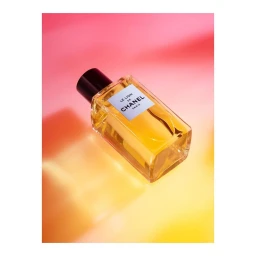 Rectangular glass perfume bottle with amber liquid and a black cap positioned diagonally on a gradient pink and yellow backdrop