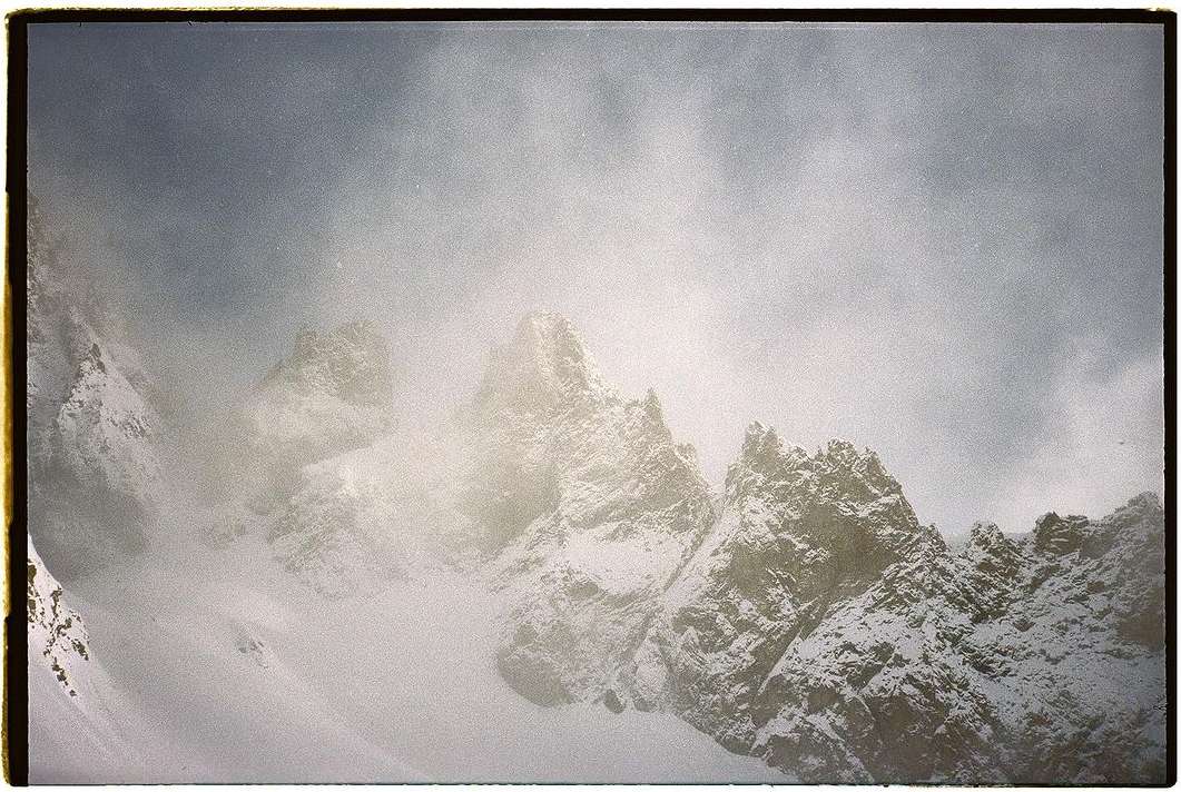 Snow-covered mountain peaks rise into a cloudy, overcast sky, creating a stark yet serene winter landscape with rugged textures.