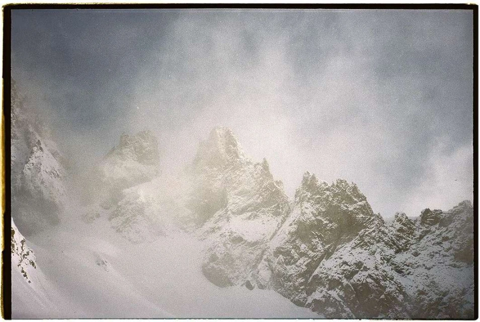 Snow-covered mountain peaks rise into a cloudy, overcast sky, creating a stark yet serene winter landscape with rugged textures.