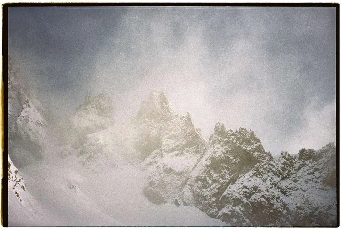 Snow-covered mountain peaks rise into a cloudy, overcast sky, creating a stark yet serene winter landscape with rugged textures.