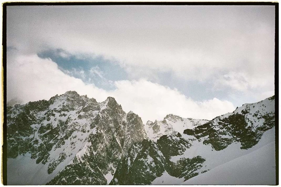 Snow-covered mountains stretch towards a cloudy sky, creating a striking landscape photograph in cool tones. The mountain range is textured.