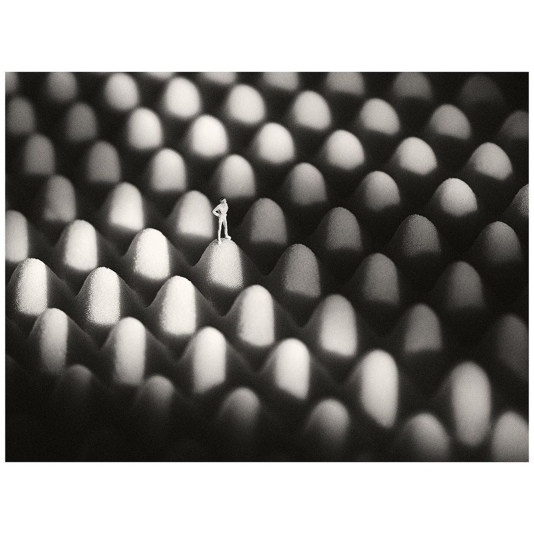 Monochrome photo of a miniature adult figure standing on patterned foam against a dark background with hard light creating strong shadows.