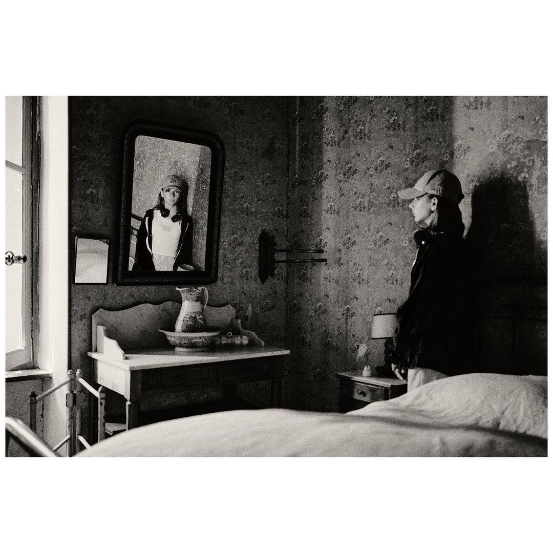 Monochrome indoor scene features an adult woman, dressed in a jacket and cap, standing in a room, with a mirror reflecting a young girl.