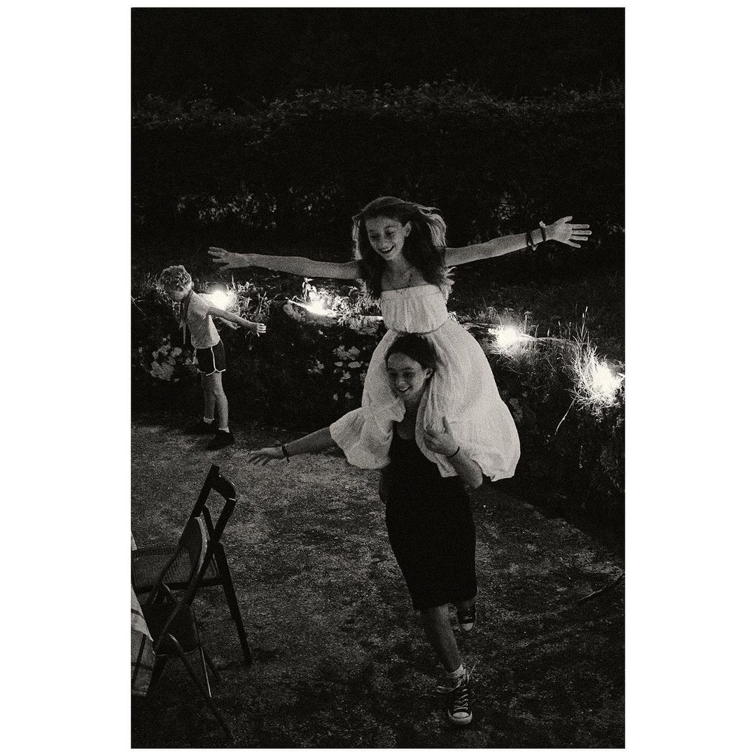 Monochrome image of a girl on another girl's shoulders, both with arms outstretched, standing in a dimly lit outdoor setting.