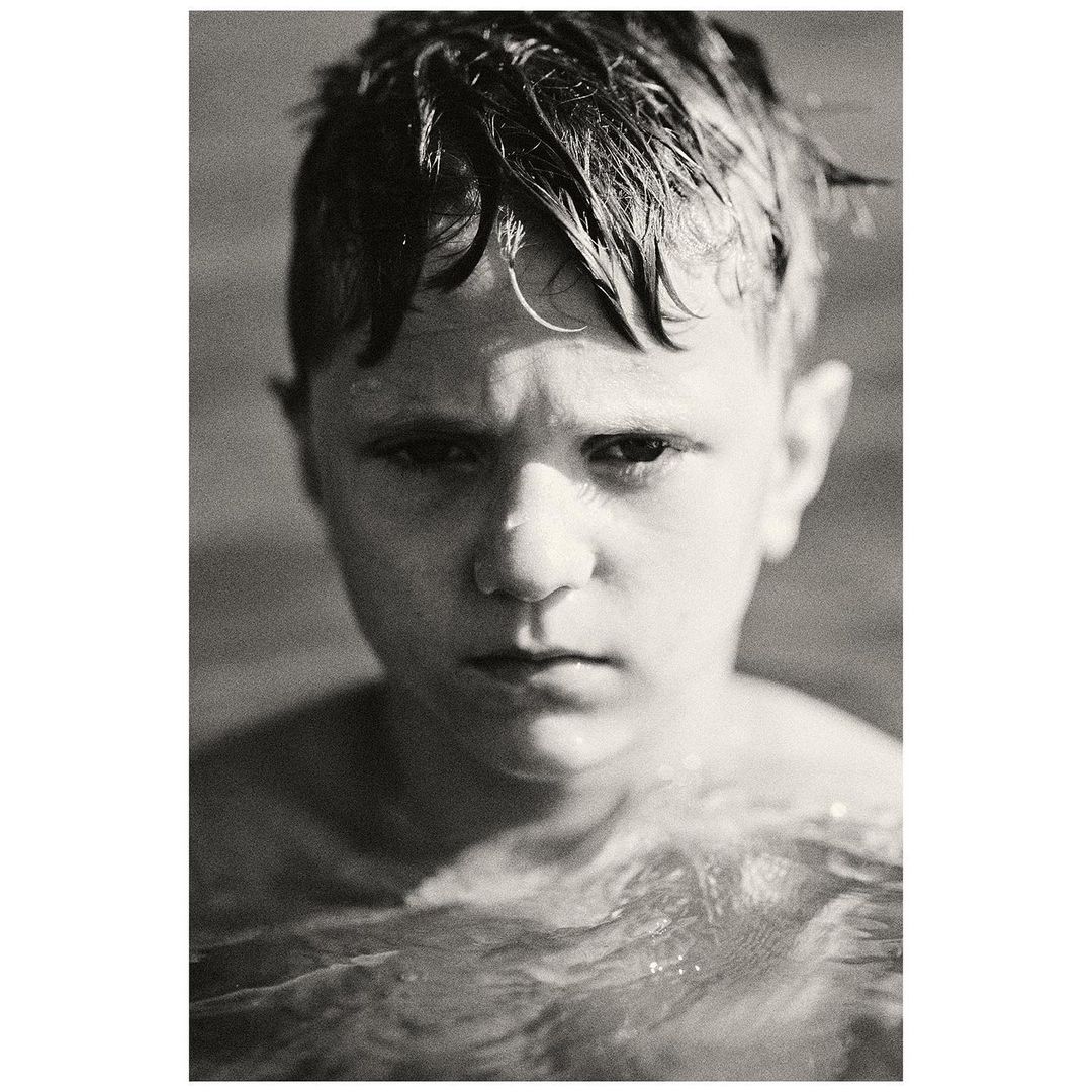 Black and white portrait of a young boy with wet hair and a serious expression, shown from the chest up and partially submerged in water.