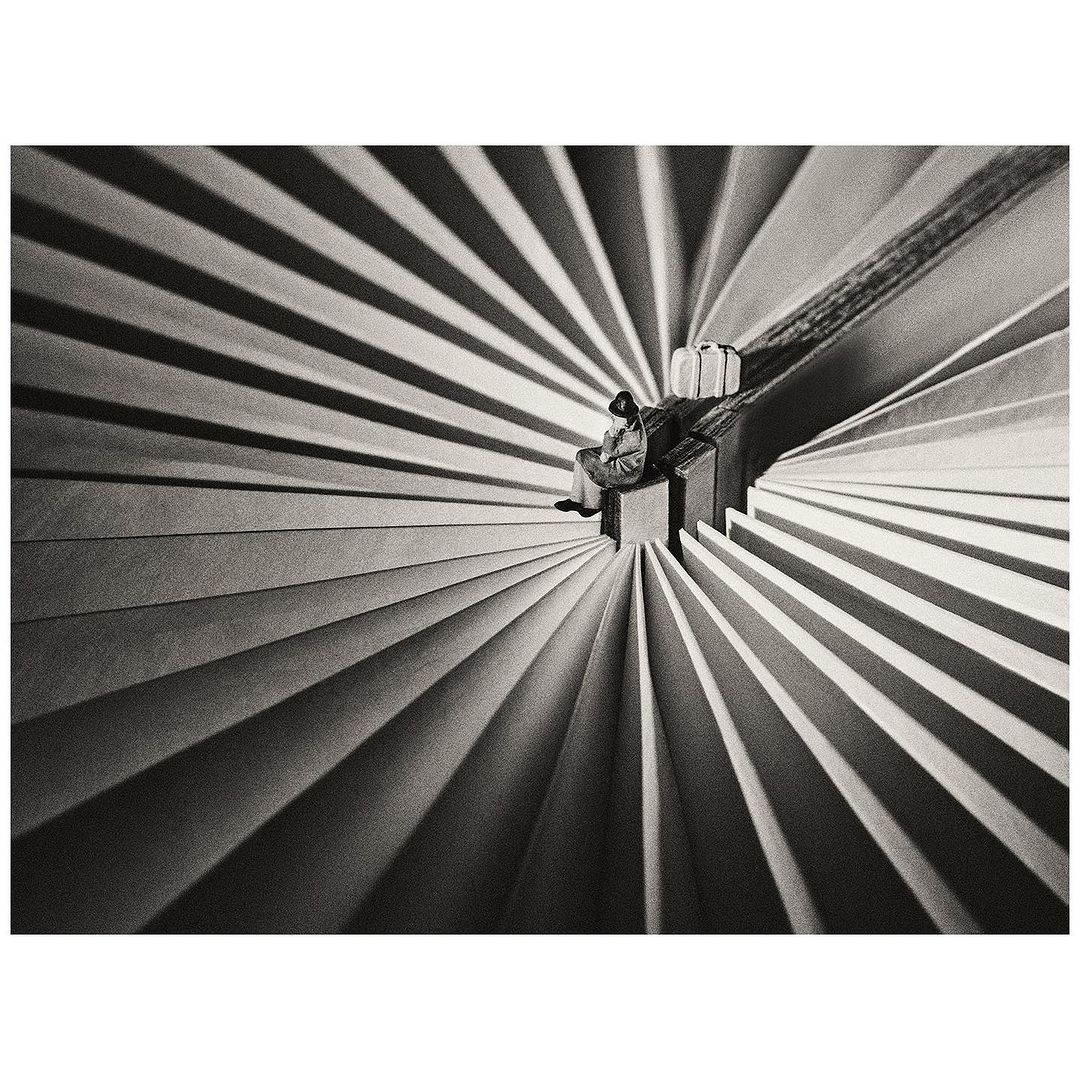 Monochrome image shows a person seated on a small platform with luggage, amidst a radiating pattern of light and shadow.