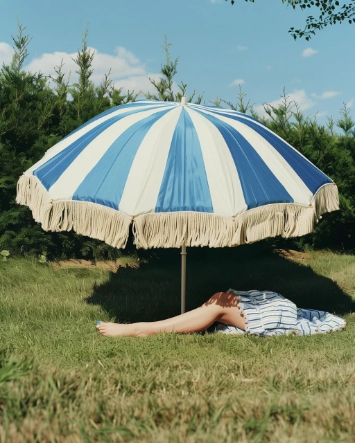 Person resting under a blue and white striped beach umbrella on green grass, surrounded by lush green trees against a clear blue sky.