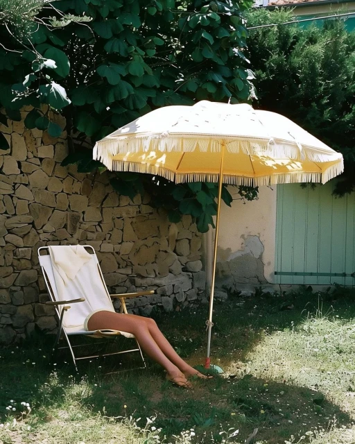 Bare legs of a woman relaxing in a lawn chair under a yellow umbrella, with a stone wall and green foliage in the background.