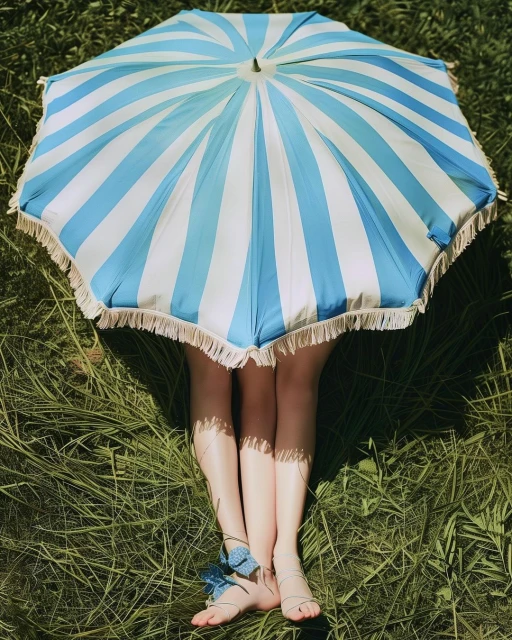 Striped blue and white umbrella casting shadow on a person laying in the green grass with ribbons tied around their ankles.