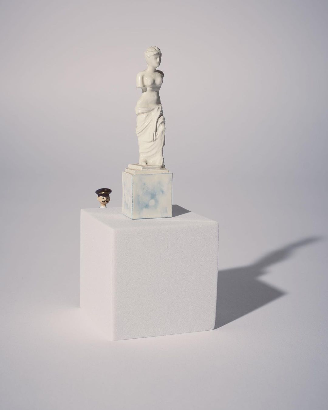 Venus de Milo statue stands atop a white cube next to a small figurine against a pale backdrop with a shadow, evoking classic art.
