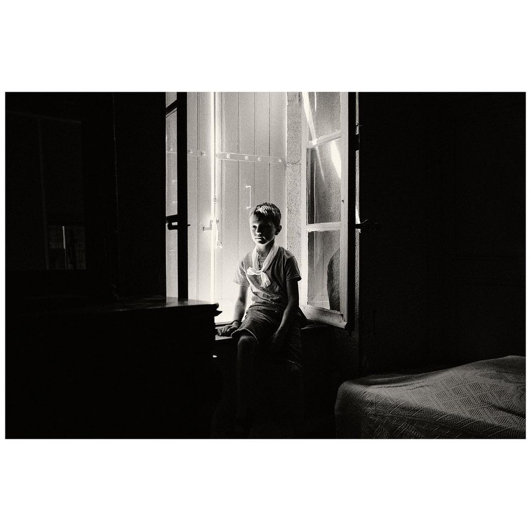 Monochrome photograph of a boy sitting by a window in a darkened room, with a bed visible in the background. Dramatic lighting.