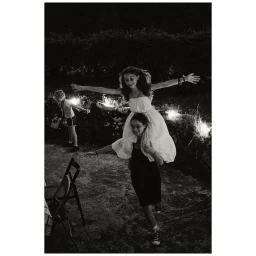 Monochrome photograph of a person carrying another person on their shoulders outdoors at night with sparklers in the background
